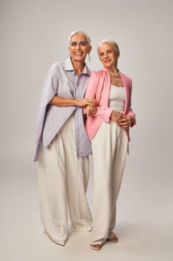 fashionable and joyful senior women in stylish casual attire smiling at camera on grey, full length clipart
