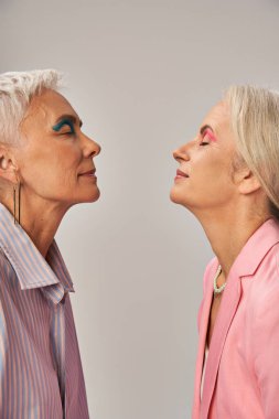 stylish senior female friends in blue and pink clothes standing with closed eyes on grey, side view clipart