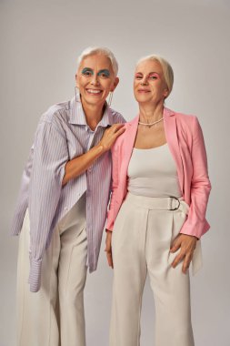 cheerful senior ladies in fashionable casual attire smiling at camera on grey, happy aging clipart