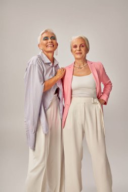 joyful senior ladies in fashionable casual attire looking at camera on grey, happy aging concept clipart