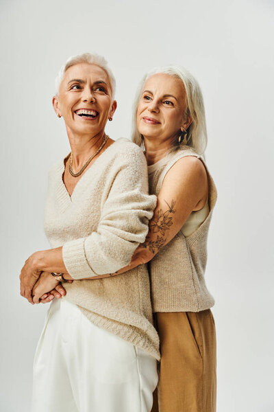 mature tattooed lady hugging joyful and stylish female friend on grey, fashionable aging concept