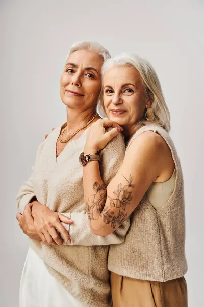 joyous senior woman with silver hair and tattoo embracing fashionable female friend on grey
