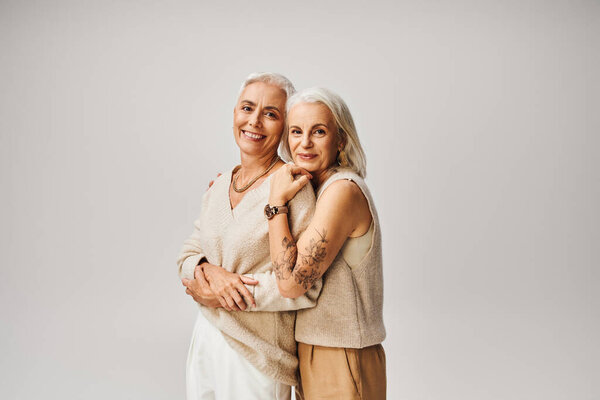 mature tattooed lady hugging cheerful and fashionable female friend on grey, positive aging