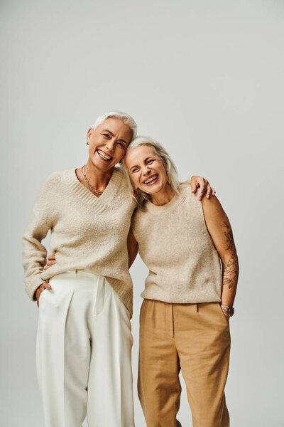 cheerful mature ladies in trendy pastel embracing and looking at camera on grey, lifelong friends