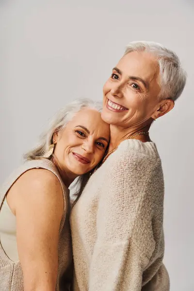 happy mature lady leaning on chest of lifelong female friend on grey, positive and stylish aging
