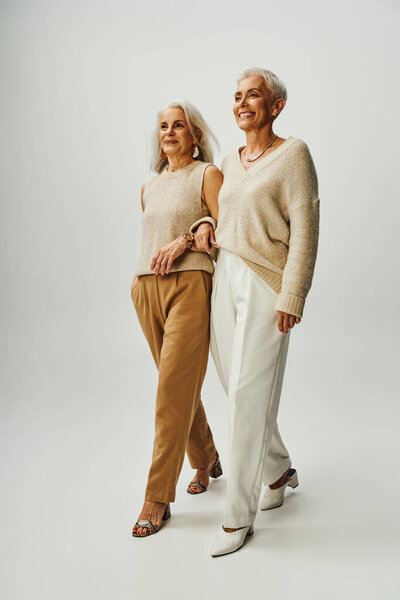 fashionable and happy senior female friends walking arm in arm on grey background, full length
