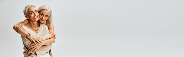 cheerful tattooed woman hugging mature and stylish female friend on grey, positive aging, banner