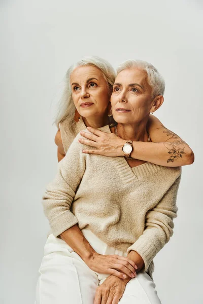 stock image senior tattooed woman embracing dreamy female friend looking away on grey, elegant lifestyle