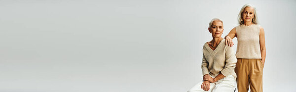 mature lady sitting near female friend standing with hand in pocket on grey, senior fashion, banner