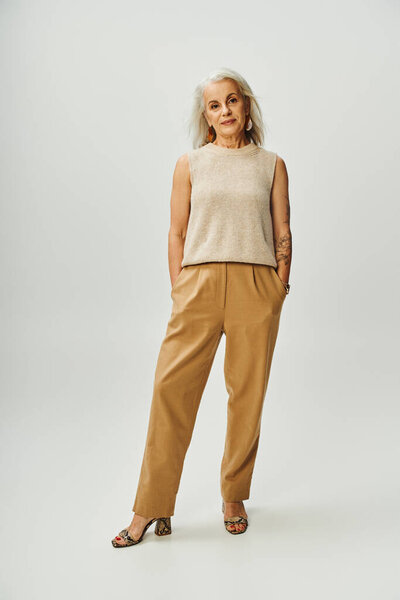 confident tattooed senior woman in stylish pastel clothes standing with hands in pockets on grey