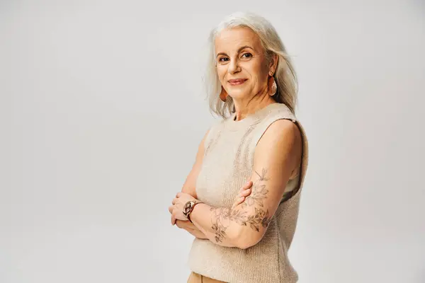 stock image tattooed mature woman in knitted west standing with folded arm and smiling on grey, age positive