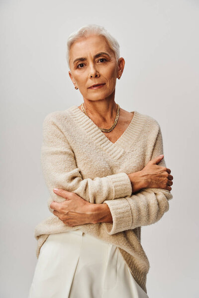 elegant mature lady in knitted jumper and golden accessories posing with folded arms on grey
