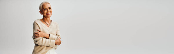 fashionable and smiley mature lady with folded arms looking at camera on grey backdrop, banner