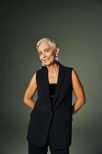 smiley senior woman with short silver hair posing in black attire with hands behind back on grey