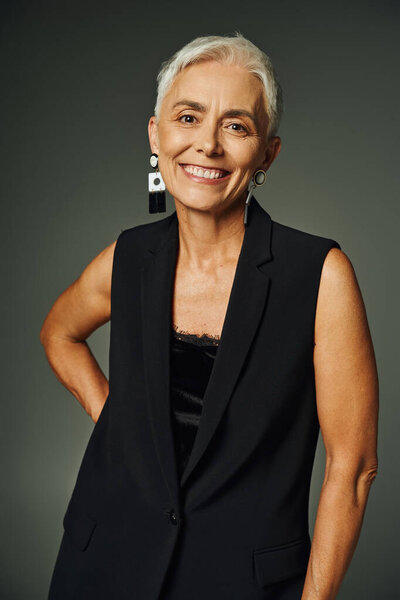 cheerful mature lady in black attire and trendy earrings looking gat camera on grey, elegant aging