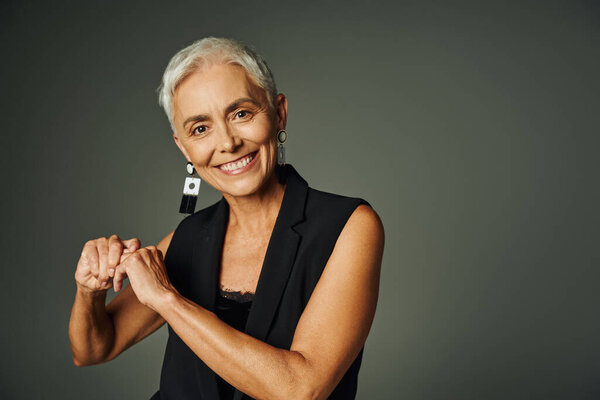 happy mature lady in stylish earrings and black attire smiling at camera on grey, elegance and charm