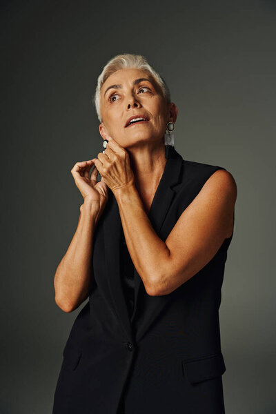 fashionable sliver haired woman in black stylish clothes adjusting earring on grey, elegance