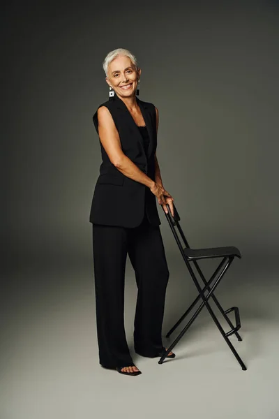 stock image full length of joyful senior lady in black classic attire posing with chair on grey, elegance