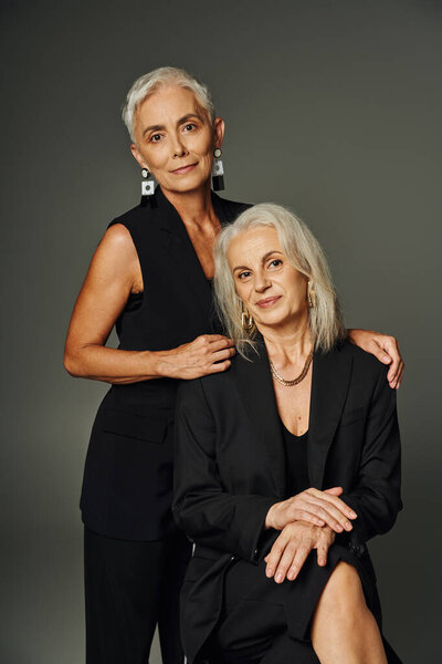 модная седовласая женщина обнимает плечи элегантной подруги, сидящей на серой