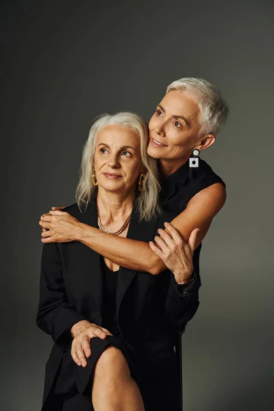 stock image dreamy senior lady in black stylish attire embracing female friend and looking away on grey