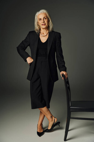full length of senior model in black attire posing with hand on hip near chair on grey, elegance