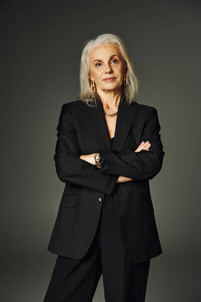 уверенная в себе зрелая дама в черном стильном одеянии позирует со сложенными руками и смотрит в камеру на сером