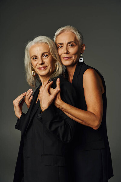 elegant mature lady in black wear hugging shoulders of female friend and looking at camera on grey