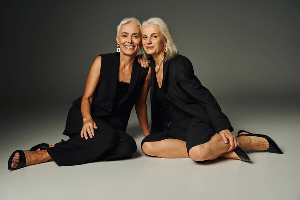 charming and happy senior models in black classic attire sitting on grey backdrop, vanity fair style