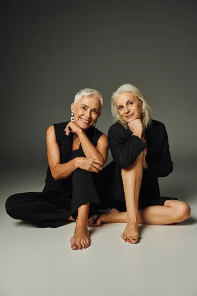 Stock image cheerful barefoot senior models in black fashionable clothes sitting and looking at camera on grey