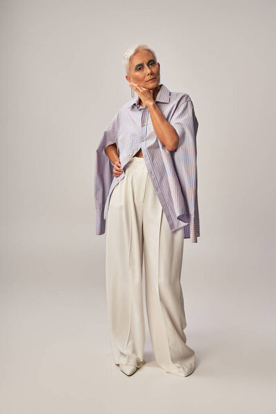 trendy aging, full length of senior woman in blue striped shirt and white pants standing on grey