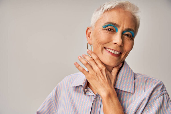 cheerful mature lady with blue eyeliner and short silver hair looking at camera on grey backdrop