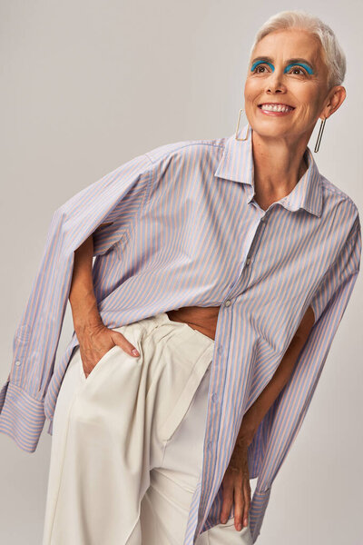 cheerful senior woman in blue striped shirt holding hand in pocket and looking away on grey