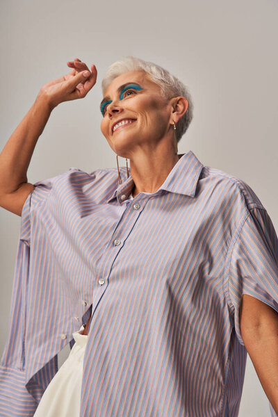 joyful and trendy senior woman with bold makeup and short silver hair looking away on grey backdrop