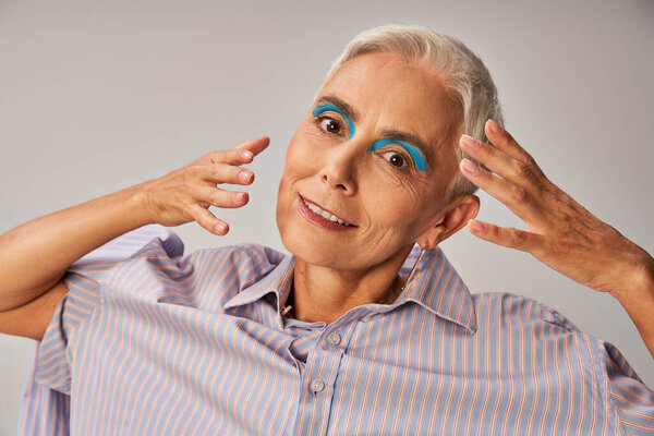 senior fashion model with blue eyeliner and short silver hair with hands near face on grey, portrait
