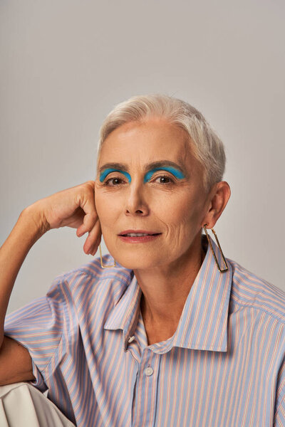 trendy senior model with short silver hair and blue eyeliner looking at camera on grey, portrait