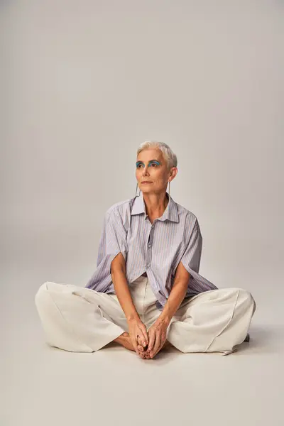 stock image pensive and barefoot mature lady in stylish casual clothes looking away while sitting on grey