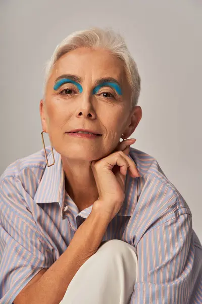 mature lady with bold makeup posing in blue striped shirt and smiling at camera on grey