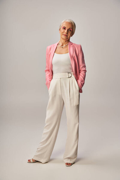 full length of senior model in pink blazer standing with hands in pockets of white pants on grey
