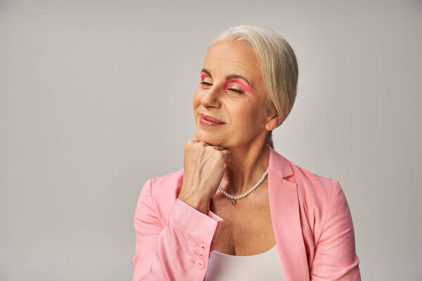 dreamy and smiling senior woman in pink blazer holding hand near and posing with closed eyes on grey
