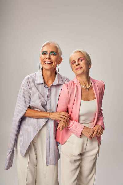 positive senior female friends in stylish casual attire smiling on grey, happy and trendy aging