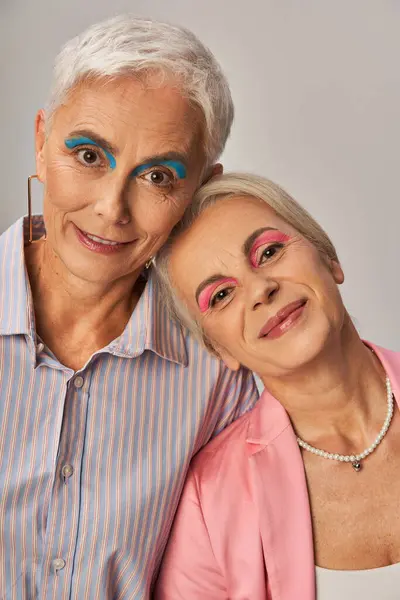 portrait of senior fashionistas with silver hair and makeup smiling at camera on grey backdrop
