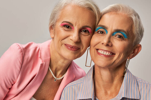 cheerful senior ladies in blue and pink clothes and makeup posing on grey, fashion and friendship