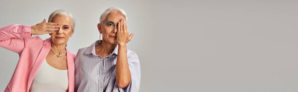 стильные зрелые женщины в синем и розовом одеянии затемняют лица руками на сером баннере