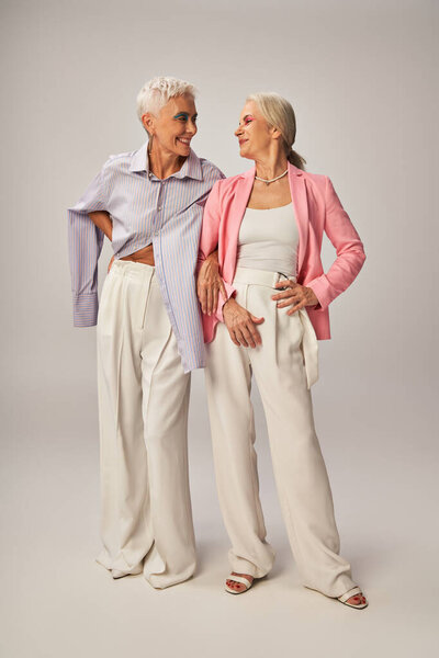 fashionable senior female friends standing with hands on hips and smiling at each other on grey