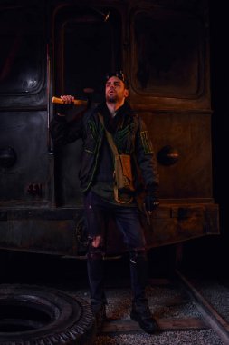 Kirli elbiseli bir adam paslı metro vagonunun yanında baltayla duruyor, felaketten sonra hayatta kalıyor.