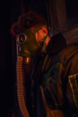 Maskeli ve ceketli bir adam karanlık yeraltı tünelinin kırmızı ışığı altında, kıyamet sonrası hayatta kalma mücadelesi veriyor.