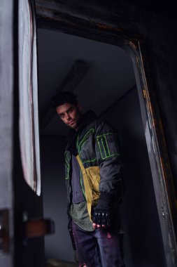 Siyah ceketli kasvetli adam terk edilmiş metro vagonunda kameraya bakıyor, kıyamet sonrası konsepti.