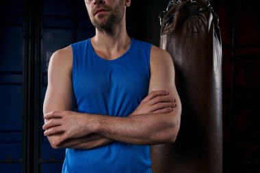 cropped view of strong man with folded arms near punching bag in dark city at night clipart