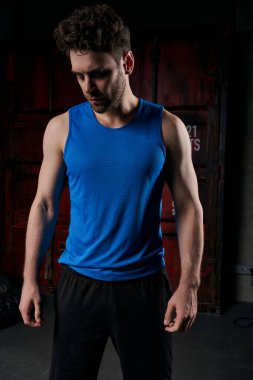 unshaven athletic man in blue tank top standing in darkness on city street at night clipart