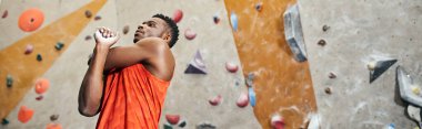 Turuncu tişörtlü Afro-Amerikan spor adamı kaya tırmanışı arka planıyla ısınıyor, afiş.